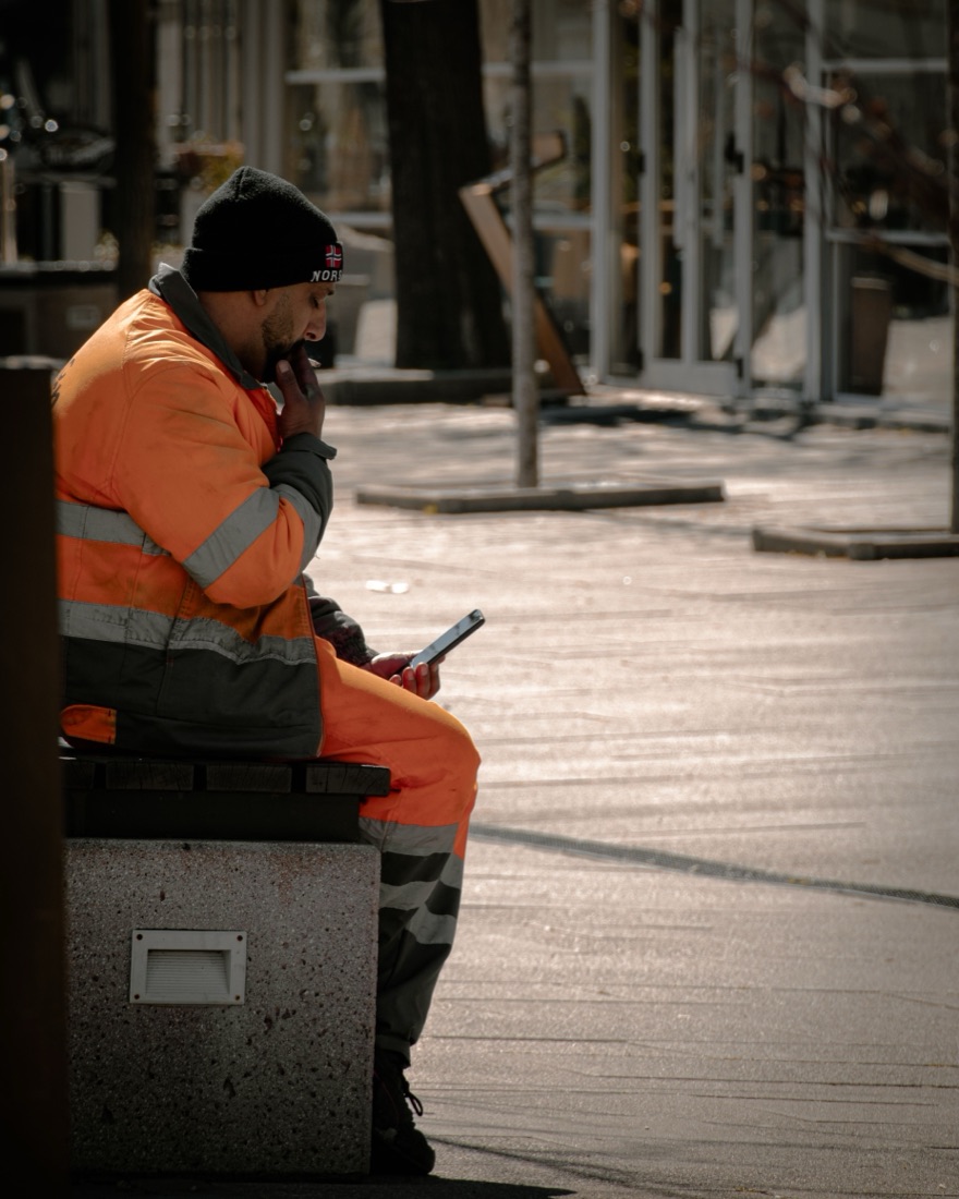 A worker in a hi-vis jacket sitting on a bench outside, reading his phone